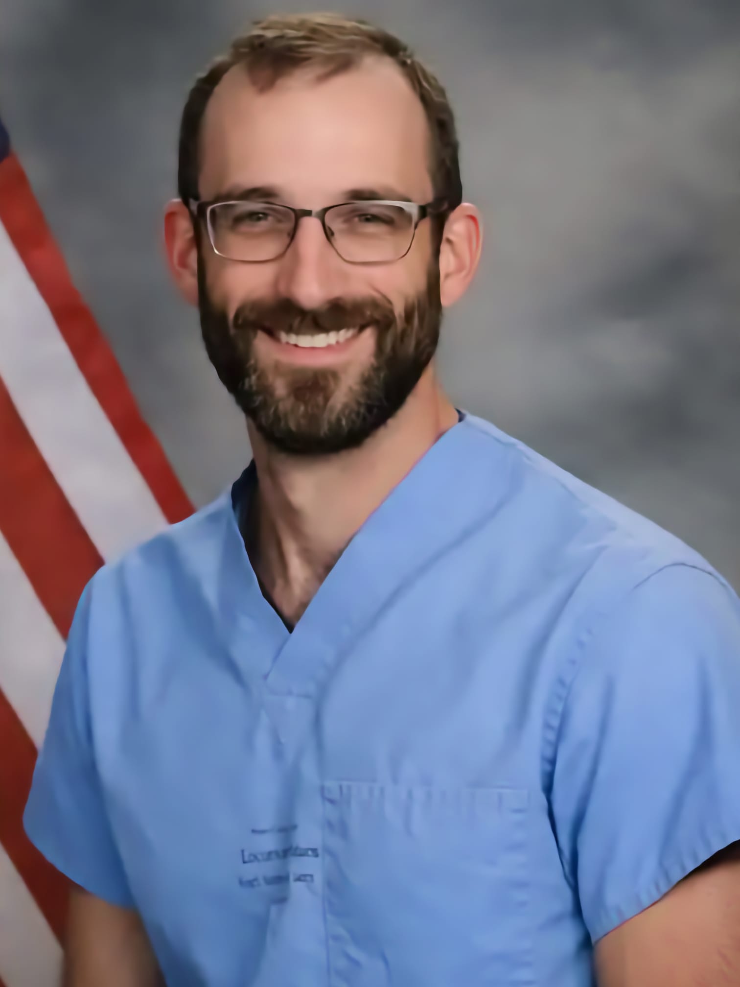 A photo of Alex Pretti, bearded white male with glasses wearing scrubs, in front of the American Flag
