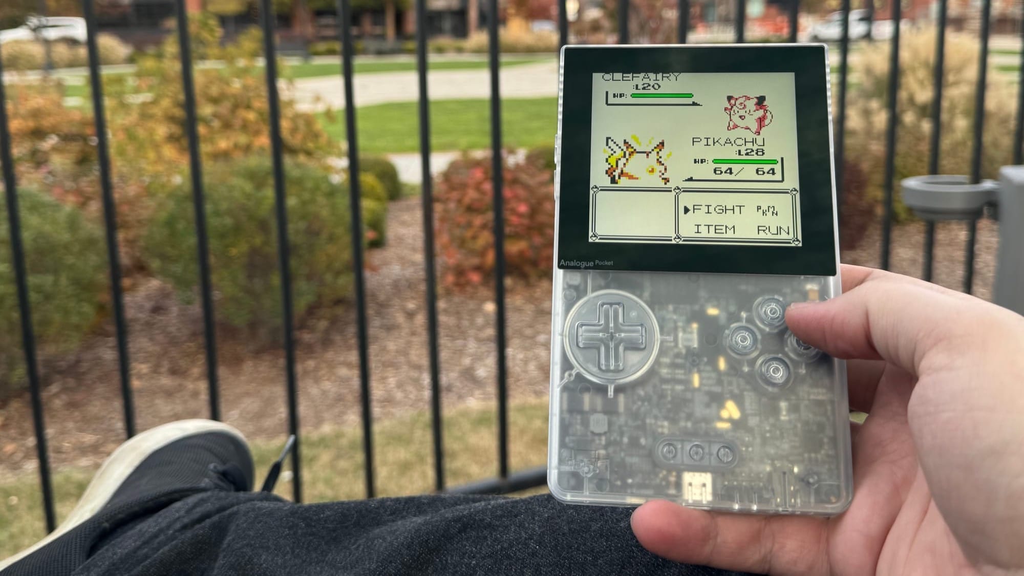 The transparent gray Analogue Pocket showing Pokemon Yellow on screen, a Pikachu fighting a Clefairy, the device held in hand and a park visible in the background.