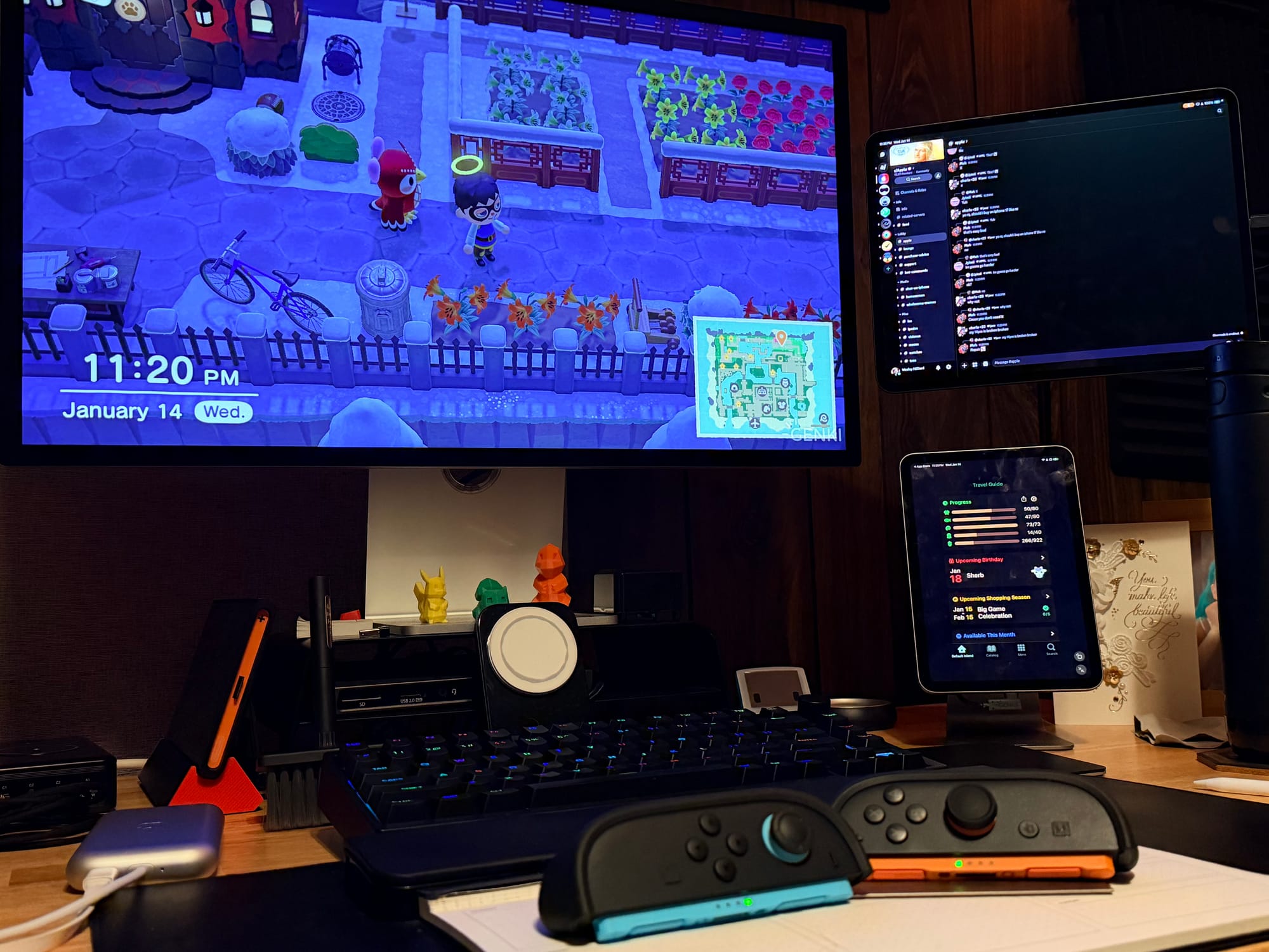A photo of a Studio Display monitor showing Animal Crossing by an iPad Pro with Discord, an iPad mini with a helper app, and two Joy Cons on the desk by a keyboard