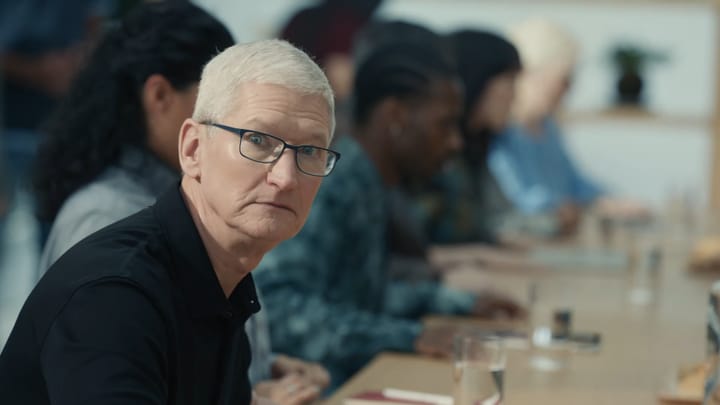 Apple CEO Tim Cook looks at the camera in shock during a skit involving mother earth, he's wearing a dark collared shirt and has white hair.