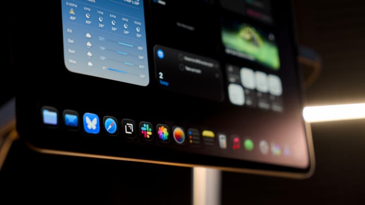 An iPad Pro on a magnetic stand shown with a desk lamp in the background, icons are visible in the dock and widgets on the Home Screen