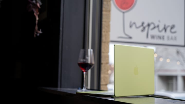 A citrus MacBook Neo on the ledge of a window sill bar by a glass of red wine, a sign for 'Inspire Wine Bar' is visible in the background